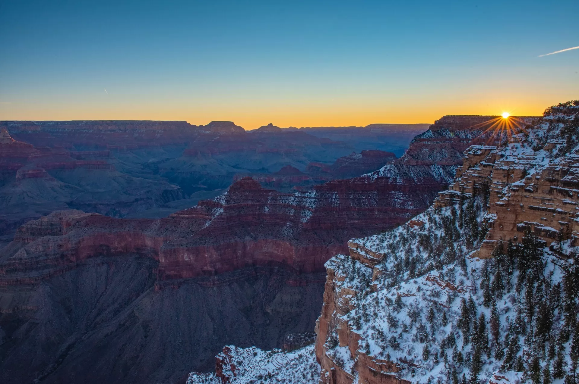 De Grand Canyon met sneeuw op de hoogste delen en de ondergaande zon op de achtergrond.