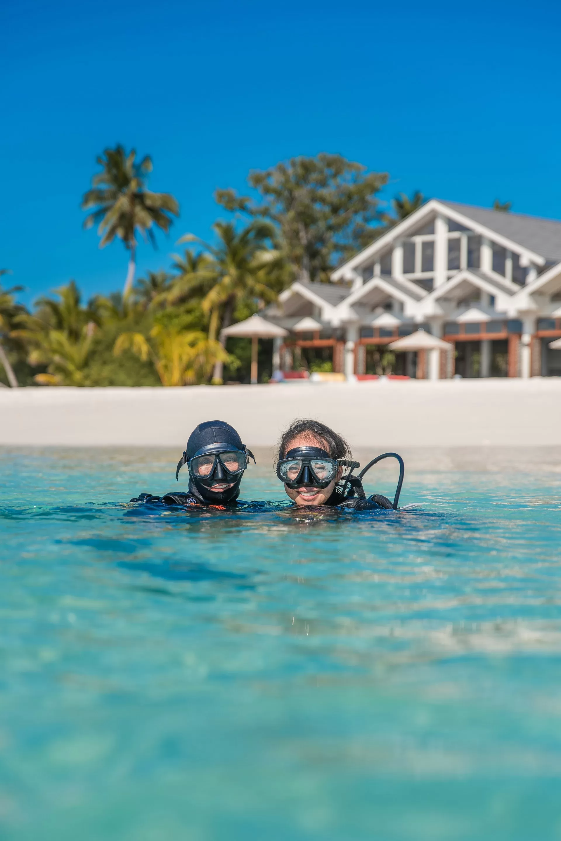 Twee snorkelaars in helder water met volledige uitrusting, tropisch strand en strandhuisje op de achtergrond zichtbaar