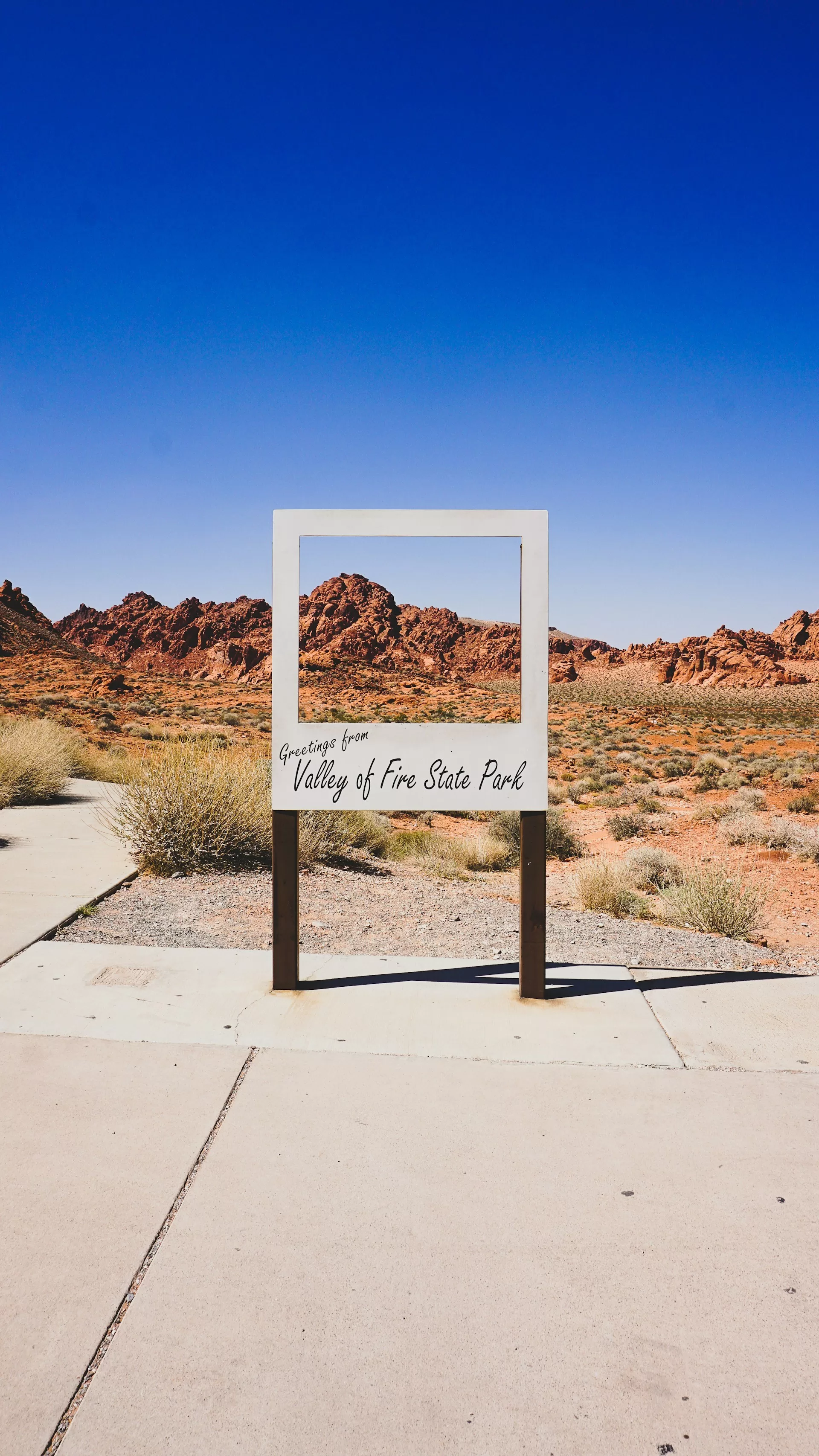 Bord met "Greetings from Valley of Fire State Park" voor rood rotslandschap en blauwe lucht in de woestijn van Nevada.