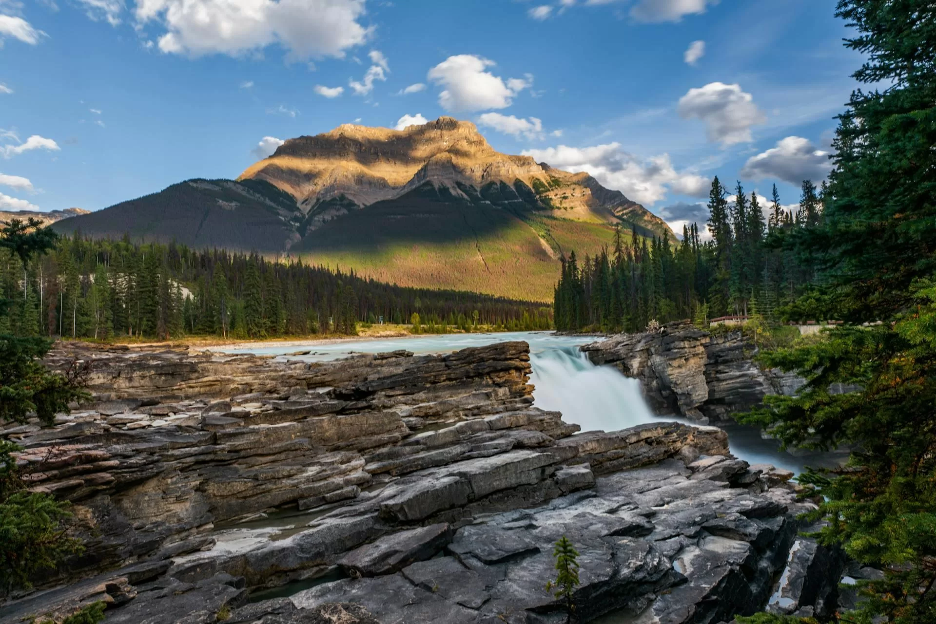 Athabasca Falls met berg op achtergrond, zonnige dag, blauwe lucht met wolken, omringd door groene dennenbomen